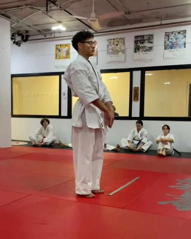 Student in a white karategi and white belt standing in a ready position on the red mats of the Nuns’ Island dojo, with other students seated along the wall