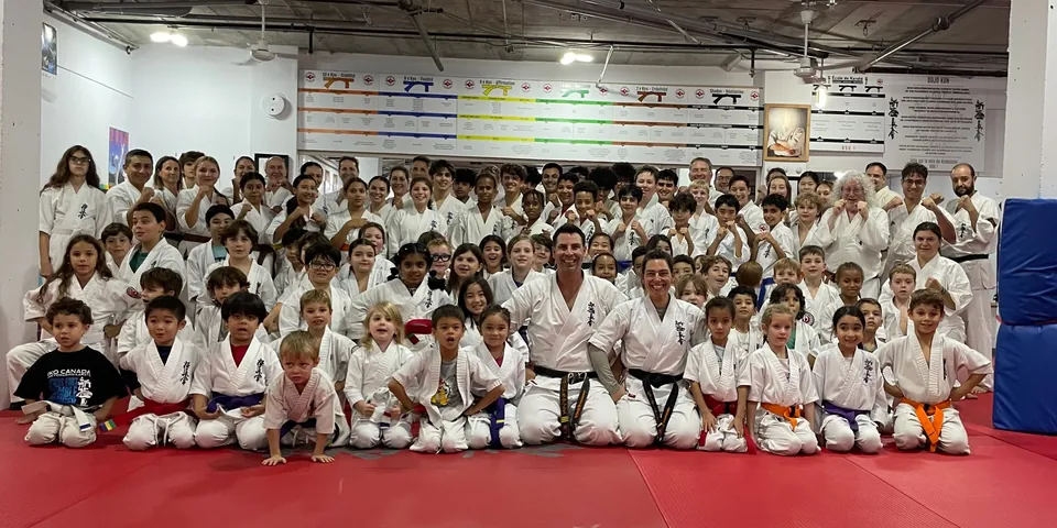 Photo de groupe au dojo Shinkyokushin de l'Île-des-Sœurs : de nombreux élèves en karatégi, enfants, adolescents et adultes, réunis sur le tatami rouge devant le tableau des ceintures et le Dojo Kun