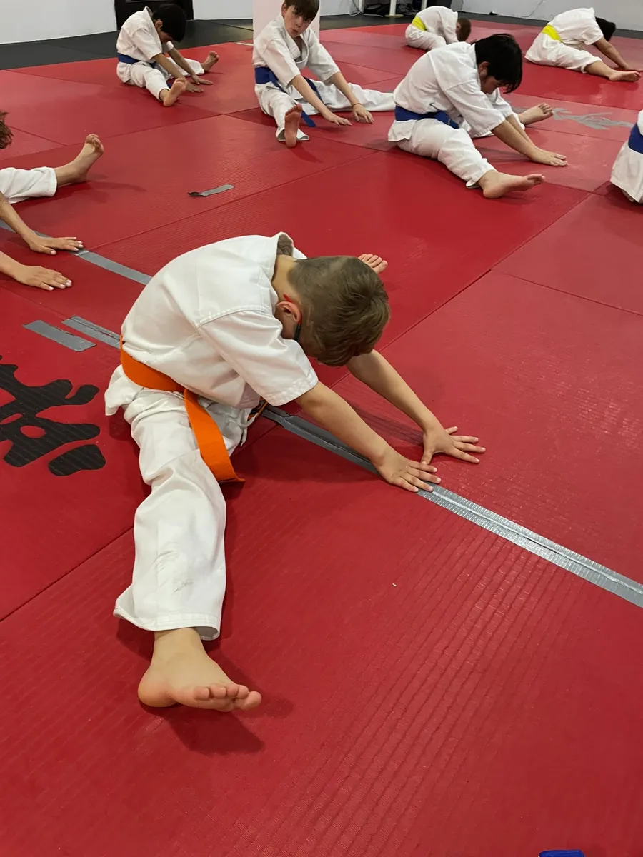 Karaté pour enfants : étirements et souplesse Karaté pour enfants : un garçon ceinture orange s'étire au sol pendant l'échauffement, entouré d'enfants ceintures bleues et jaunes, sur les tapis rouges du dojo de l'Île-des-Sœurs.