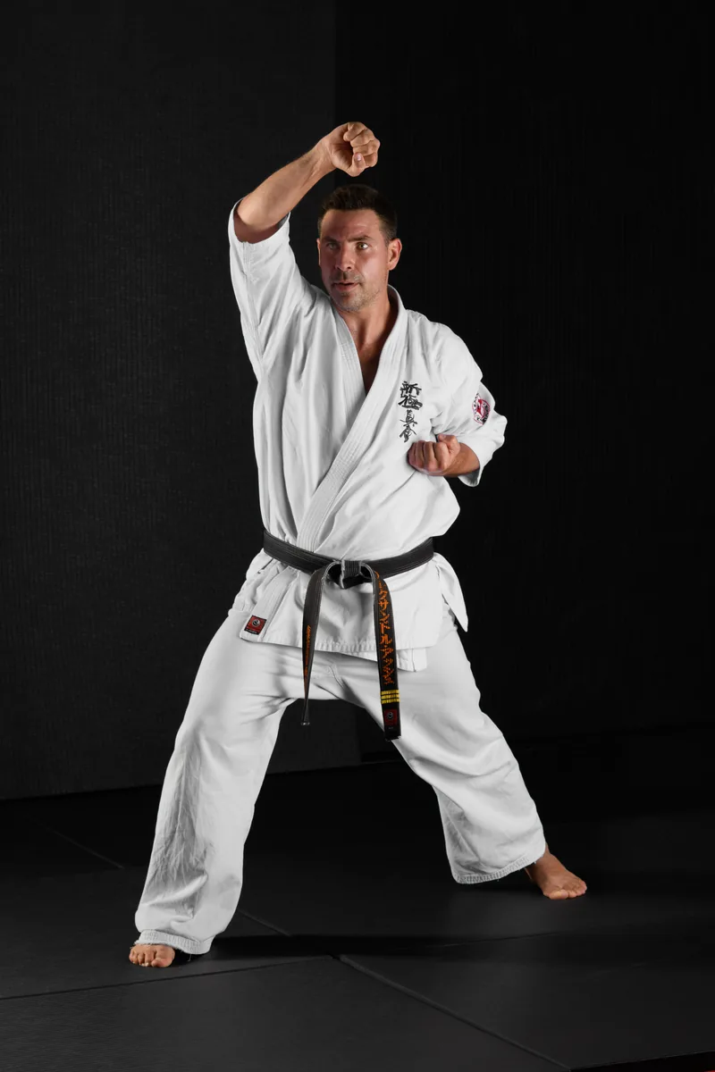 Sensei Alexandre Haché : posture de karaté Kyokushin Sensei Alexandre Haché en posture de karaté Kyokushin, le bras droit levé, en dogi blanc et ceinture noire brodée à son nom.