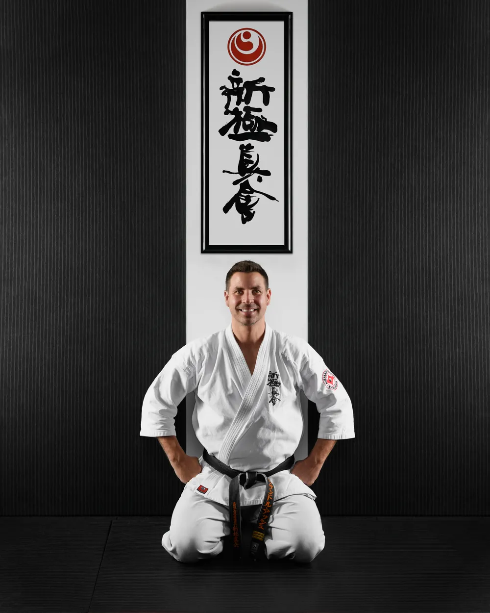 Sensei Alexandre Haché, ceinture noire, assis en seiza au dojo Shinkyokushin Karaté Île-des-Sœurs, sous la calligraphie Shinkyokushinkai